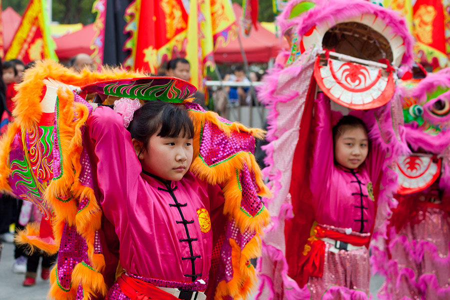 Dziewczynki w pochodzie z okazji karnawału. Dzieci są ubrane w różowe tradycyjne stroje chińskie. Ciągną za sobą dekoracje, które powiewają na powietrzu. Wokół nich tłum ludzi.