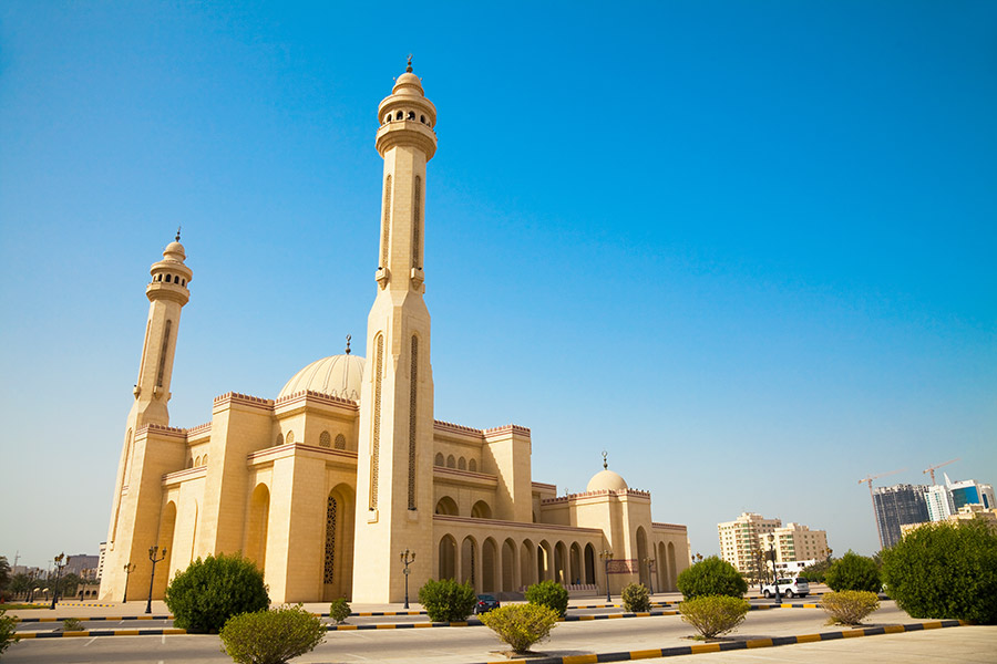 Meczet w Manamie, stolicy Bahrajnu widoczny od boku. Po lewej dwie wysokie i wąskie wieże. Na środku kopuła. Przed budynkiem rosną niskie krzewy.