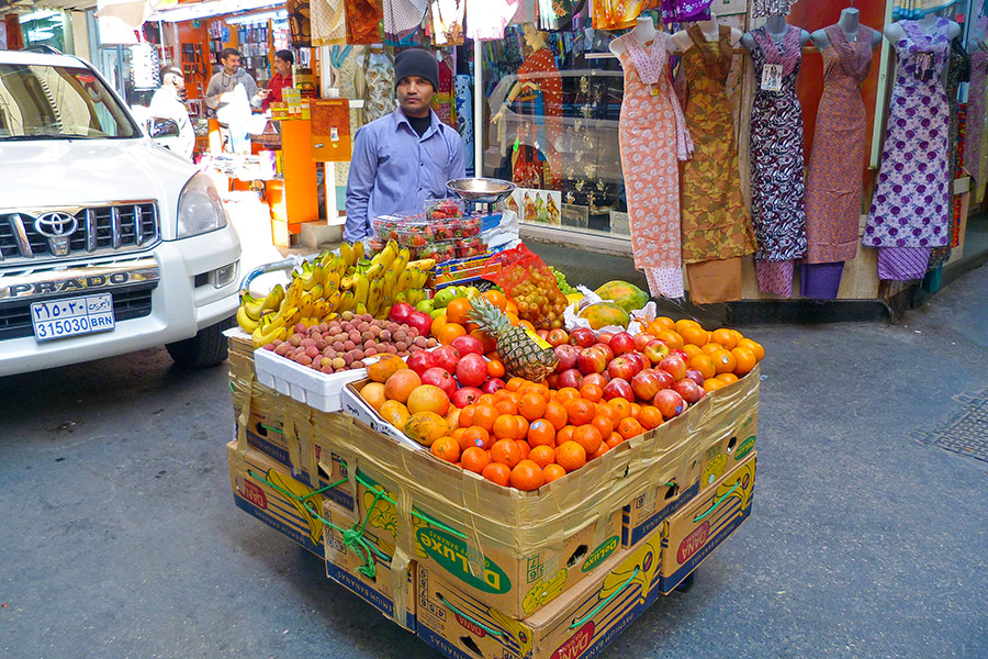 Mieszkaniec w drodze na targ. Wiezie kartony z owocami: banany, ananasy, pomarańcze, granaty. Za nim wystawa z odzieżą oraz samochód.