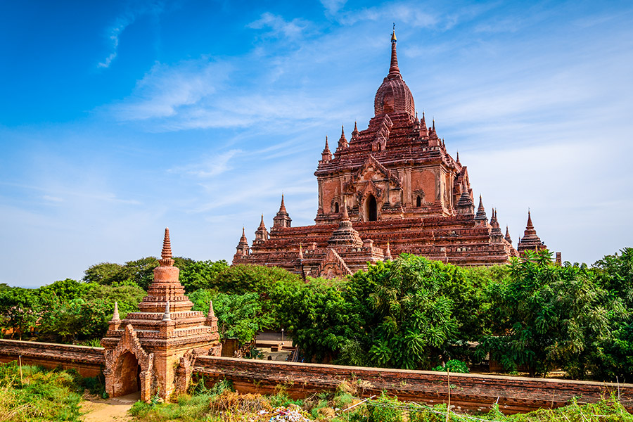Świątynia buddyjska w Bagan. Świątynia ma kilka kondygnacji i liczne wieże. Do świątyni prowadzi brama zakończona wieżą. Pomiędzy bramą a świątynią rośnie dużo drzew.